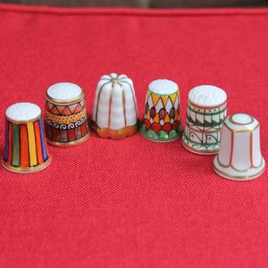 Pode incluir: Uma coleção de seis dedais de cerâmica dispostos em semicírculo. Cada dedal apresenta um design único, incluindo riscas verticais, padrões geométricos e motivos florais em cores como laranja, verde, vermelho e detalhes em dourado.
