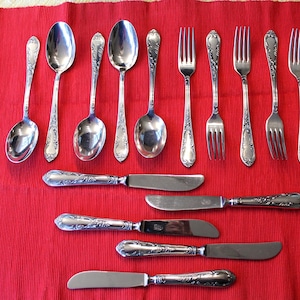 Può includere: Un assortimento di posate antiche, tra cui cucchiai, forchette e coltelli, disposte su una tovaglia rossa. Le posate hanno manici ornati e dettagliati e una superficie lucida. Gli utensili sono di varie dimensioni.