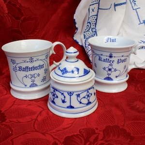 Puede incluir: Un juego de tres tazas de café de porcelana blanca con diseños florales azules y blancos. Las tazas están etiquetadas como "Kaffeebecher", "Kaffee Pott" y "Flut".