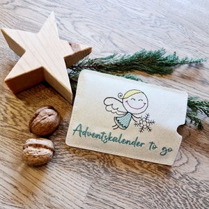 May include: A white fabric advent calendar with a green embroidered angel holding a snowflake. The text "Adventkalender to go" is embroidered in green below the angel.
