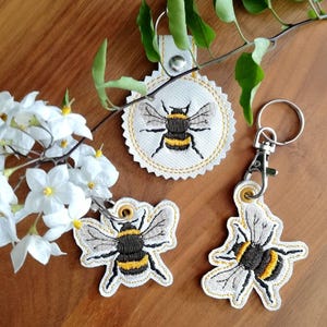 Puede incluir: Tres llaveros bordados con forma de abeja. Los llaveros presentan un fondo blanco con un borde cosido amarillo y un diseño detallado de abeja en negro, amarillo y gris. Un llavero es redondo y dos tienen forma de abeja.