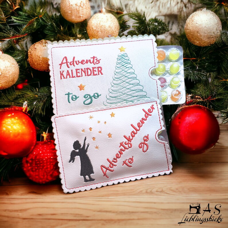 May include: Two white felt pouches with embroidered Christmas designs. The top pouch has a green Christmas tree and the text "Advent Kalender to go". The bottom pouch has a silhouette of an angel and the text "Adventskalender to go".