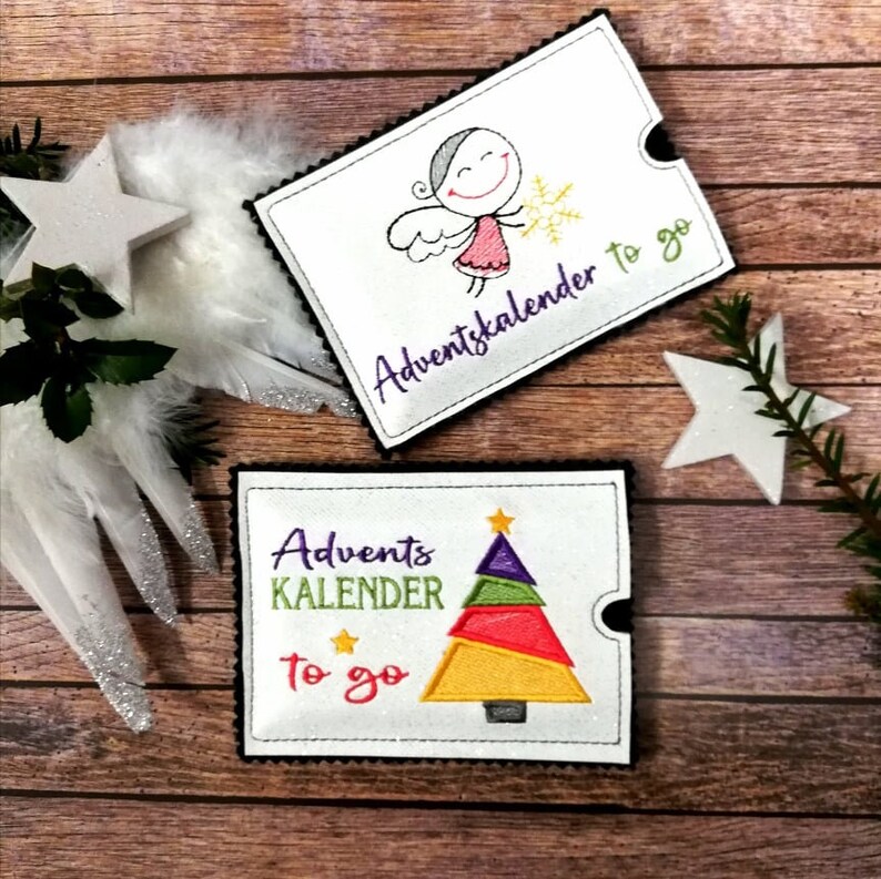 May include: Two white fabric gift card holders with black stitching. One features an angel with the text "Adventkalender to go". The other features a colorful Christmas tree with the text "Advent KALENDER to go".