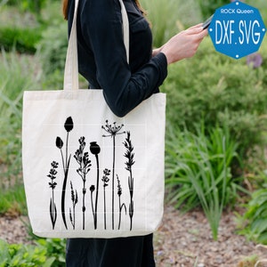 May include: A white tote bag featuring a black silhouette of assorted wildflowers and grasses printed on the front. The bag is being held by a person.