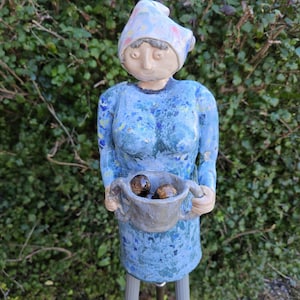 Puede incluir: Estatua de jardín de cerámica caprichosa de una mujer con vestido y sombrero azules, sosteniendo un cuenco con dos objetos marrones. La estatua tiene un acabado azul moteado y está montada sobre patas grises. El fondo es un seto verde.
