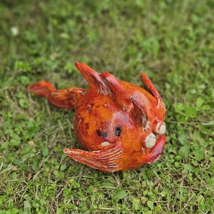 Può includere: Una scultura artigianale in ceramica arancione e rossa di una creatura fantastica con orecchie a punta, una lunga coda e piccoli occhi rotondi. La scultura è adagiata sull'erba verde, mettendo in risalto la sua superficie strutturata e il design unico.