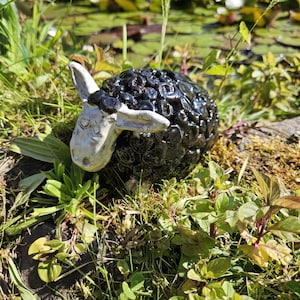 Könnte beinhalten: Eine Keramik-Schafskulptur mit einem schwarzen, strukturierten Körper und einem weißen Gesicht und Ohren. Das Schaf befindet sich inmitten von grünem Gras und Laub, mit einem Teich im Hintergrund. Die Skulptur hat eine glänzende Oberfläche.