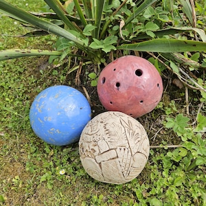 Op de afbeelding: Drie decoratieve keramische bollen in een tuin. Eén is blauw, één is rood met gaten en de derde is beige met een geëtst ontwerp. De bollen liggen op groen gras en zijn omgeven door bladeren.