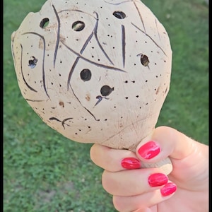 Peut inclure: Un objet creux, arrondi et de couleur beige clair, doté d'un manche, présentant une série de perforations circulaires et des marques linéaires incisées sombres sur sa surface. L'objet a un aspect organique et texturé avec une finition lisse.