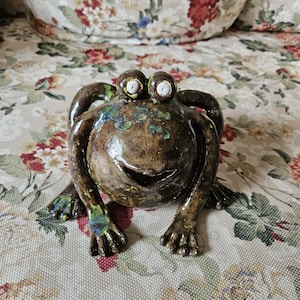 Könnte beinhalten: Eine Keramikfrosch-Skulptur mit glänzend brauner Oberfläche. Der Frosch hat große, ausdrucksstarke Augen und einen lächelnden Mund. Grüne und blaue Akzente sind am Körper und an den Füßen zu sehen. Die Skulptur steht auf einer Oberfläche mit floralem Muster.
