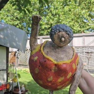 May include: A whimsical ceramic sculpture of a woman in a red and yellow polka-dot swimsuit, with one arm raised in a fist. The figure has a gray, textured hairstyle and a neutral skin tone. The sculpture is held up by a wooden stick.