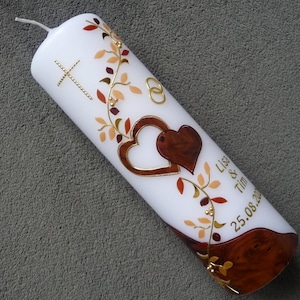 Könnte beinhalten: Weiße Stumpenkerze verziert mit einem goldenen Kreuz, zwei Herzen und floralen Akzenten in Braun, Gold und Orange. Die Kerze hat einen braunen Sockel und die Namen "Lisa & Tim" sowie das Datum "25.08.20".