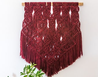May include: A burgundy macrame wall hanging with a geometric diamond pattern and fringe. The textile is suspended from a natural wood dowel, creating a decorative and textured wall accent. The wall hanging is a unique piece of home decor.