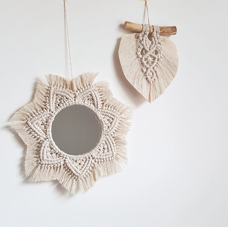May include: Two macrame wall hangings. One is a starburst-shaped mirror with a circular mirror in the center. The other is a leaf-shaped macrame piece attached to a wooden dowel. Both are made of cream-colored cotton cord and are hanging on a white wall.