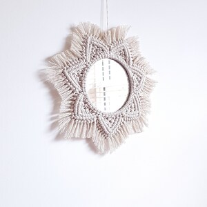 May include: A round mirror with a cream-colored macrame frame. The frame has a starburst design with knotted details and fringe. The mirror is hanging against a white wall.