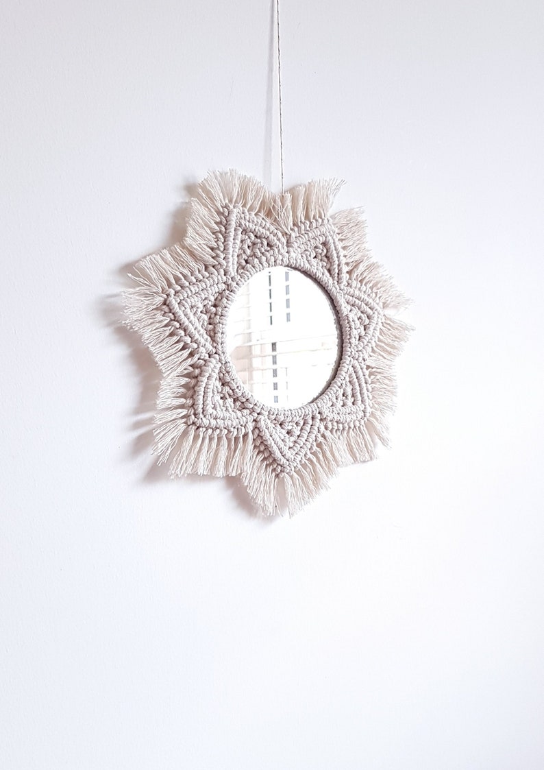 May include: A round mirror with a cream-colored macrame frame. The frame has a starburst design with knotted details and fringe. The mirror is hanging against a white wall.