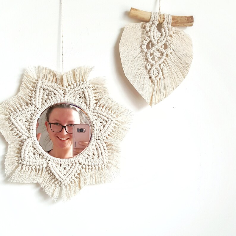May include: Two macrame wall hangings. One is a starburst-shaped mirror with a cream-colored fringe. The other is a leaf-shaped wall hanging with a wooden branch at the top. Both are made of cream-colored macrame.