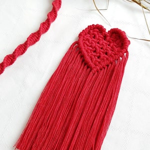 Può includere: Un cuore in macramè rosso fatto a mano con lunghe frange e un cordoncino ritorto. Il cuore è lavorato all'uncinetto con un motivo strutturato. La frangia pende sotto il cuore. Il cordoncino è ritorto a spirale. Gli oggetti sono su una superficie bianca.