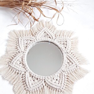 May include: A round mirror with a cream-colored macrame sunburst design. The macrame has a detailed woven pattern and fringe. Dried brown grasses are in the background. This decorative mirror is a bohemian style home decor item.