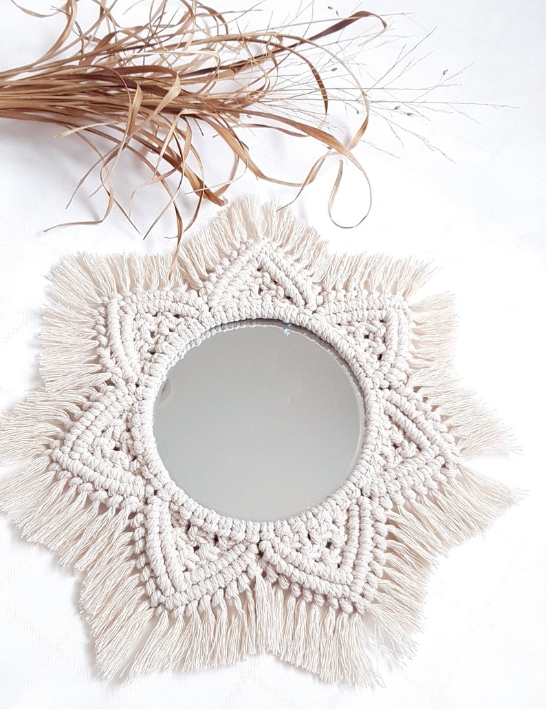 May include: A round mirror with a cream-colored macrame sunburst design. The macrame has a detailed woven pattern and fringe. Dried brown grasses are in the background. This decorative mirror is a bohemian style home decor item.