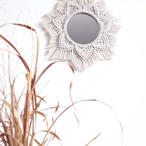 May include: A round mirror with a cream-colored macrame sunburst design. The mirror is hanging on a white wall. The macrame has a geometric pattern and fringe. Dried ornamental grass is in the foreground.