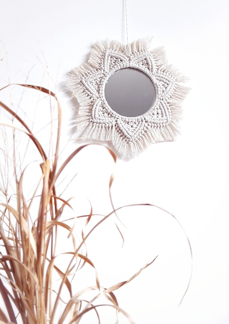 May include: A round mirror with a cream-colored macrame sunburst design. The mirror is hanging on a white wall. The macrame has a geometric pattern and fringe. Dried ornamental grass is in the foreground.