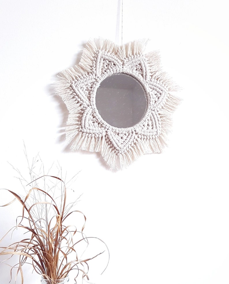 May include: A round mirror with a cream-colored macrame sunburst design. The mirror is centered within the macrame, and the design features intricate knotting and fringe details. The mirror is hanging against a white wall.
