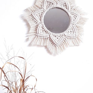 May include: A round mirror with a cream-colored macrame sunburst design. The mirror is centered within the macrame, and the design features intricate knotting and fringe details. The mirror hangs against a white wall.