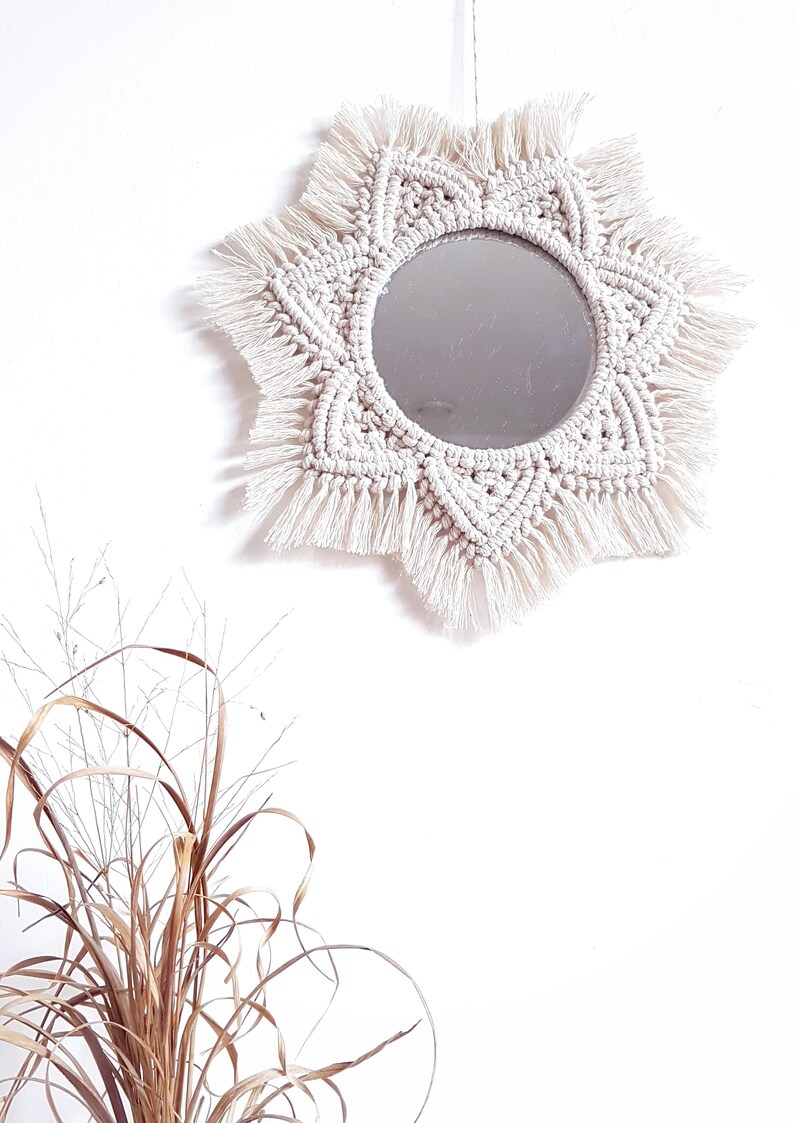 May include: A round mirror with a cream-colored macrame sunburst design. The mirror is centered within the macrame, and the design features intricate knotting and fringe details. The mirror hangs against a white wall.