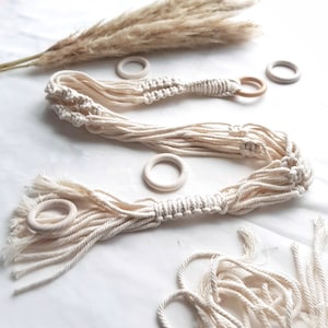 May include: Cream-colored macrame plant hangers with wooden rings. The hangers are made of twisted cotton cord and are designed to hold potted plants. Dried pampas grass is in the background. The macrame plant hangers are a decorative item.