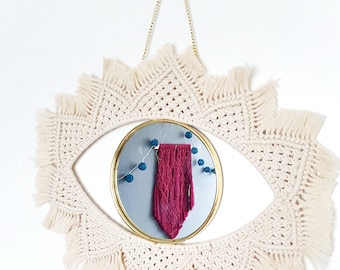 May include: A decorative wall mirror with a macrame eye-shaped frame in cream color. The mirror has a gold-colored frame and a burgundy macrame piece hanging inside. Small blue pom-poms are strung above the burgundy piece.