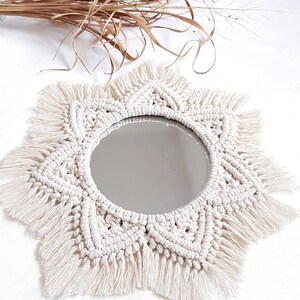 May include: A round mirror with a cream-colored macrame sunburst design. The mirror has a fringe around the edge and is surrounded by a woven pattern. Dried plants are in the background.