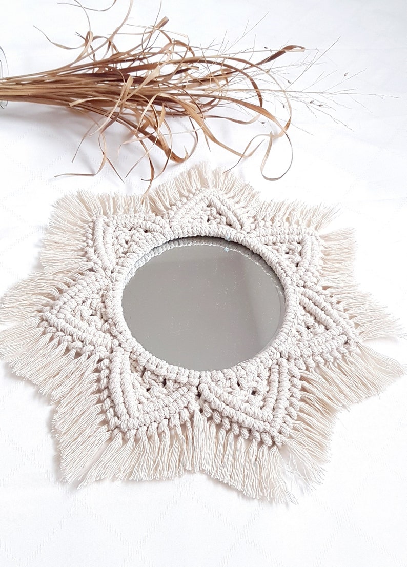 May include: A round mirror with a cream-colored macrame sunburst design. The mirror has a fringe around the edge and is surrounded by a woven pattern. Dried plants are in the background.