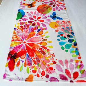 May include: A white table runner with a colourful watercolour floral design. The design features pink, red, orange, yellow, green, blue, and purple flowers with butterfly accents.