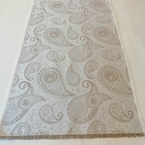 Puede incluir: Un camino de mesa blanco con un patrón de paisley en color marrón claro. El camino de mesa tiene un borde deshilachado.