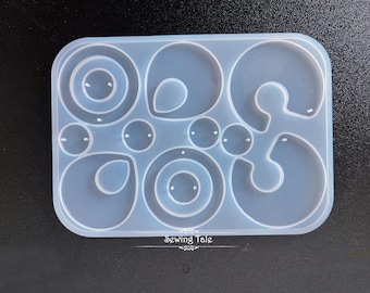 Puede incluir: Un molde de silicona transparente y rectangular con varias formas de pendientes, incluyendo círculos, gotas y diseños curvos. El molde tiene una superficie lisa y está diseñado para la elaboración de joyas. El texto "Sewing Tale" está en la parte inferior.