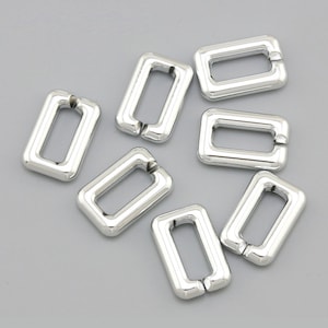 Puede incluir: Un conjunto de ocho anillos de metal plateados, cada uno con forma rectangular y una abertura redondeada. Los anillos están dispuestos de forma dispersa sobre una superficie gris.