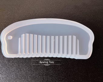 May include: A translucent, light blue comb-shaped mold with a rounded handle and a series of vertical teeth. The mold has a scalloped edge and the text "Sewing Tale" is visible at the bottom. The comb mold is likely for crafting or resin projects.