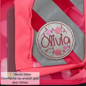 Könnte beinhalten: Ein leuchtend pinker Rucksack mit einem runden silbernen Glitzeraufnäher, der den Namen Olivia in pinker Schreibschrift zeigt. Der Rucksack hat diagonale graue Reflexstreifen, einen pinken Reißverschluss und den Textüberlagerungshinweis Neues Silber Oberfläche rau anstatt glatt aber Glitzer.