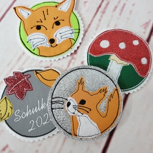 Könnte beinhalten: Vier Filzapplikationen mit gestickten Designs. Ein roter und weißer Pilz, ein Fuchs mit orangem Fell, ein Eichhörnchen mit orangem Fell und ein grauer Kreis mit dem Text "Schule 2022".
