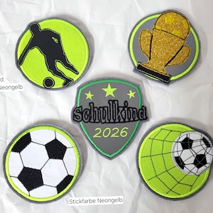 Könnte beinhalten: Fünf bestickte Aufnäher mit Fußballmotiven. Einer zeigt die schwarze Silhouette eines Spielers, einer einen goldenen Glitzerpokal, einer einen klassischen schwarz-weißen Fußball, einer einen Ball im Netz und ein Schild mit dem Text Schulkina 2026.