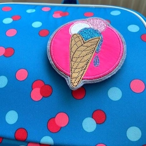 Könnte beinhalten: Ein kreisförmiger, bestickter Aufnäher mit einer beigen Waffel und blau-rosa glitzernden Eiskugeln auf leuchtend pinkem Hintergrund. Der Aufnäher ist auf einem blauen Stoff mit roten, pinken und hellblauen Punkten befestigt.