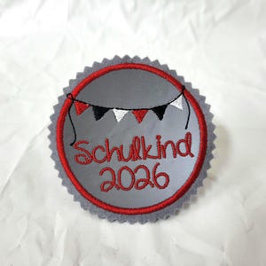 Könnte beinhalten: Ein kreisförmiger, grauer Stickaufnäher mit gezacktem Rand. Das Design zeigt eine rote Kreisumrandung, eine Girlande aus roten, schwarzen und weißen dreieckigen Wimpeln sowie den roten Sticktext Schulkind 2026 in der Mitte.
