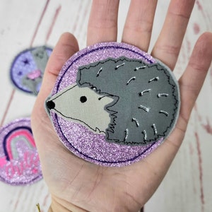 Könnte beinhalten: Ein kreisförmiger bestickter Aufnäher mit einem grauen Igel auf glitzerndem violettem Hintergrund. Der Igel hat ein hellgraues Gesicht und einen dunkelgrauen Körper mit gestickten Details. Zwei weitere runde Aufnäher sind im Hintergrund zu sehen.