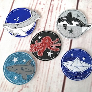 Könnte beinhalten: Fünf runde Filzpatches mit verschiedenen Designs zum Thema Meer. Jeder Patch hat einen weißen Umriss und einen Glitzer-Hintergrund. Die Designs umfassen einen Wal, einen Oktopus, einen Hai, eine Orca und ein Papierboot.