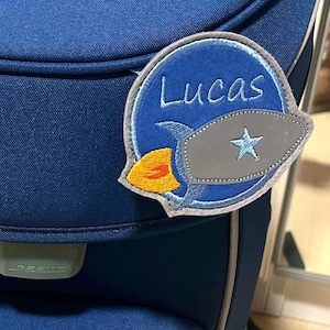 Könnte beinhalten: Ein kreisförmiger, bestickter Aufnäher auf einer dunkelblauen Tasche. Der Aufnäher hat einen blauen Hintergrund mit dem in Weiß gestickten Namen Lucas, eine graue Rakete mit einem hellblauen Stern und eine gelbe Flamme am unteren Rand.