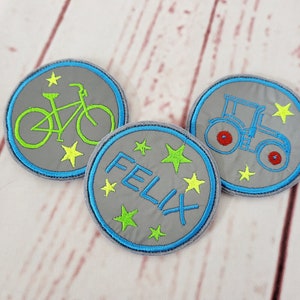 Könnte beinhalten: Drei graue Filzpatches mit blauem Rand. Ein Patch hat ein grünes Fahrrad mit gelben Sternen, einer hat einen blauen Traktor mit roten Rädern und gelben Sternen und einer hat den Namen "Felix" in blau mit gelben Sternen.