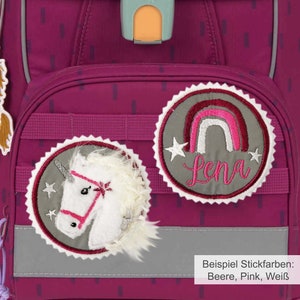 Könnte beinhalten: Ein rosa Rucksack mit zwei gestickten Patches. Ein Patch zeigt ein Einhorn mit einem rosafarbenen Zaumzeug und einer weißen Mähne. Der andere Patch zeigt einen Regenbogen mit einem weißen Stern auf jeder Seite und dem Namen "Lena" in rosa gestickt. Der Text "Beispiel Stickfarben: Beere, Pink, Weiß" ist unter den Patches sichtbar.