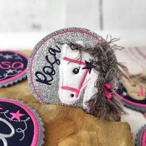 Könnte beinhalten: Ein kreisförmiger, glitzernder Aufnäher mit einem weißen Pferdekopf mit rosa Zaumzeug und einem dunkelblauen Stern. Der Name "Rosa" ist in dunkelblauer Schrift gestickt. Weitere Aufnäher mit ähnlichen Designs sind sichtbar.