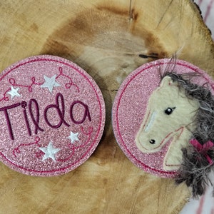 Könnte beinhalten: Zwei kreisförmige Aufnäher mit rosa Glitzer-Finish. Ein Aufnäher zeigt den Namen Tilda in dunkelviolettem Garn gestickt, umgeben von weißen Sternen. Der andere zeigt einen cremefarbenen Pferdekopf mit einer Mähne aus dunkelgrauem Kunstfell.